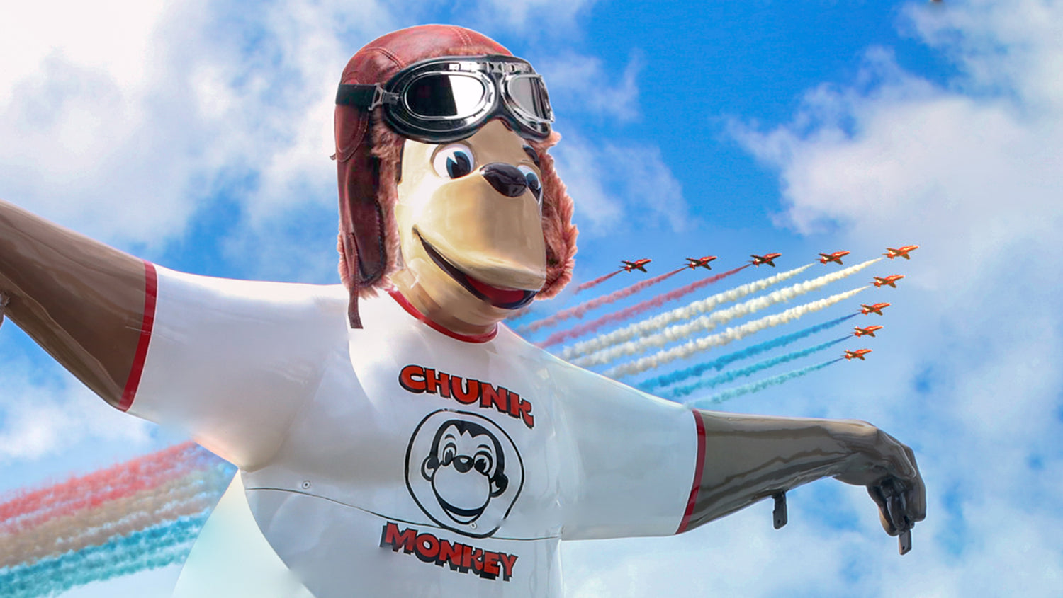 The Chunk Monkey mascot wearing a pilots hat and goggles with the red arrows flying in the sky behind him