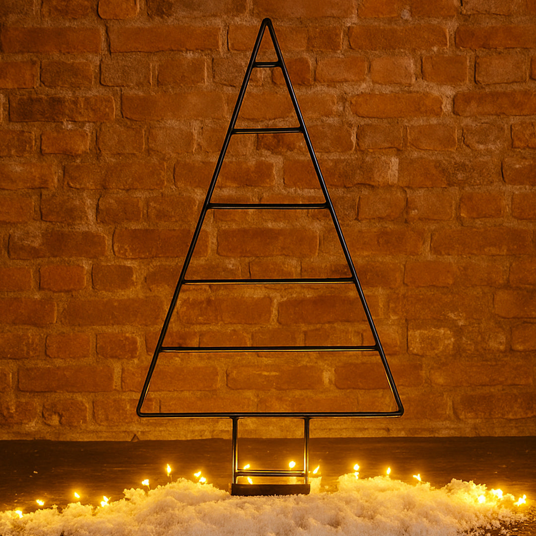 Black metal Christmas tree decoration against a brick wall with lights on the ground.