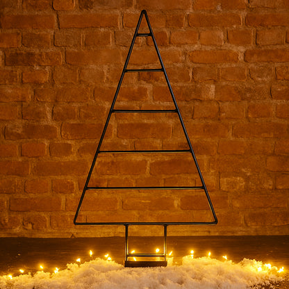 Black metal Christmas tree decoration against a brick wall with lights on the ground.
